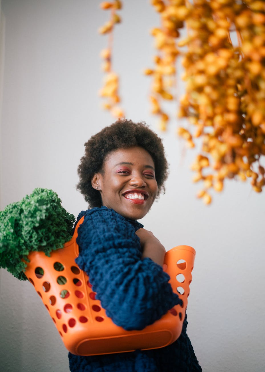 happy young black woman holding basket with lettuce on shoulder and cluster of yellow dates in grocery store on gray background