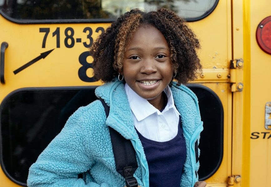 happy black schoolgirl with backpack standing near school bus